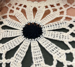 A close-up of a crocheted lace doily with a black center and white radiating petals, laid flat on a dark green fabric surface.