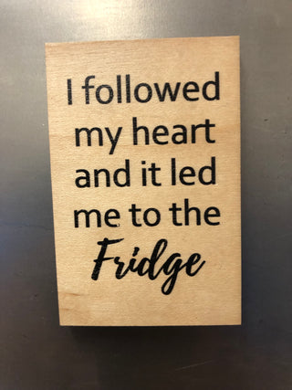 A rectangular wooden magnet with black printed text that reads, 'I followed my heart and it led me to the Fridge,' photographed on a dark gray surface.