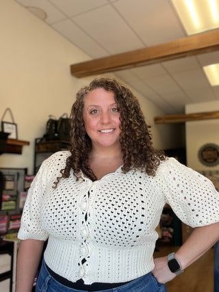 A smiling woman with curly brown hair wearing a white open-knit short-sleeve cardigan top and blue jeans, standing in what appears to be a boutique interior.