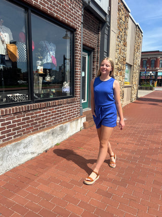 A young woman walking on a red brick sidewalk next to a brick and stone storefront, wearing a bright blue sleeveless romper and tan sandals.