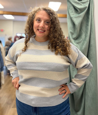 A woman with curly brown hair wearing a cream and light blue-gray striped knit sweater and blue jeans, standing in a fitting room or boutique.