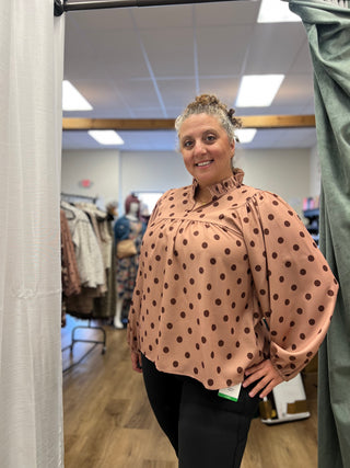 A woman wearing a peach-colored long-sleeve blouse with dark brown polka dots and black pants, standing in a fitting room or boutique.
