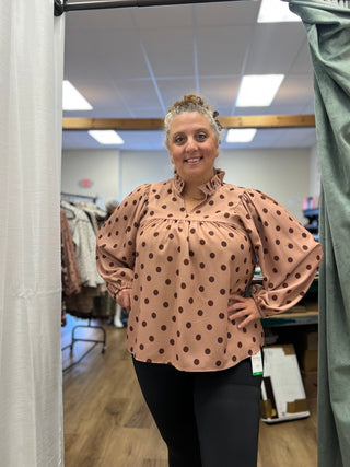 A woman modeling a peach-colored blouse with dark brown polka dots and voluminous puff sleeves, standing in a fitting room.