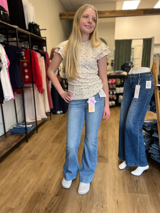 A young blonde woman modeling light-wash blue flare jeans in a clothing store, wearing a cream floral t-shirt and white sneakers. A mannequin displaying dark blue flare jeans stands to her right.