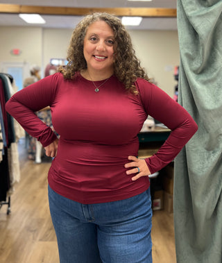 A woman with curly brown hair wearing a fitted, long-sleeve burgundy top and blue jeans, standing in a retail store environment.