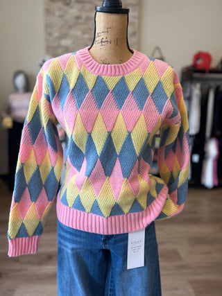 A multicolor knit sweater with a pink, yellow, and blue harlequin diamond pattern, displayed on a mannequin torso wearing blue jeans in a clothing store.