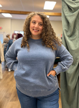 A woman with curly brown hair wearing a heathered slate blue crewneck sweatshirt and blue jeans, standing in a retail store fitting area.