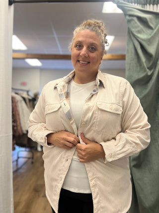 A woman wearing a light beige corduroy button-up shirt over a white t-shirt, standing in a fitting room or boutique.