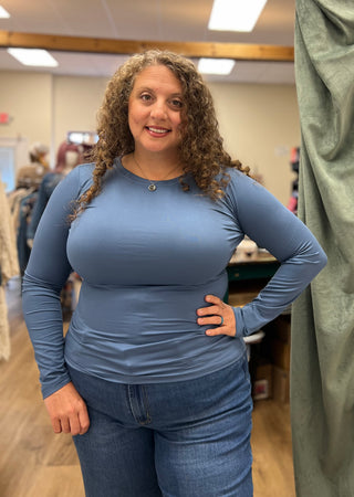 A woman with curly brown hair wearing a fitted, long-sleeve dusty blue top and blue jeans, standing in a retail store environment.