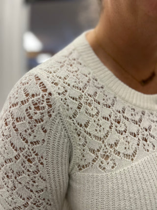 Close-up of a person wearing a cream-colored open-knit sweater with a detailed lace pattern across the chest and shoulder.