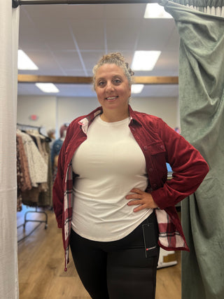 A woman posing in a fitting room wearing a white crewneck t-shirt, black leggings, and a red maroon button-up flannel shirt jacket.