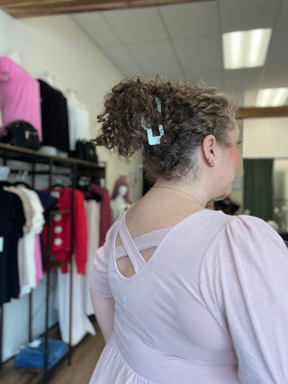 A woman with curly brown hair wearing a light lavender dress, seen from behind in a clothing store. She is wearing a light blue hair clip to hold her hair up.