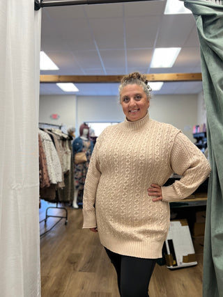 A woman wearing a cream cable-knit mock-neck sweater dress stands in a fitting room, with clothing racks and store fixtures visible in the background.