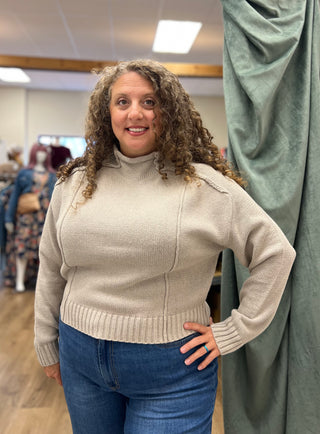 A woman with curly brown hair wearing a beige ribbed mock-neck sweater and blue jeans, standing in a fitting room or boutique next to a green curtain.
