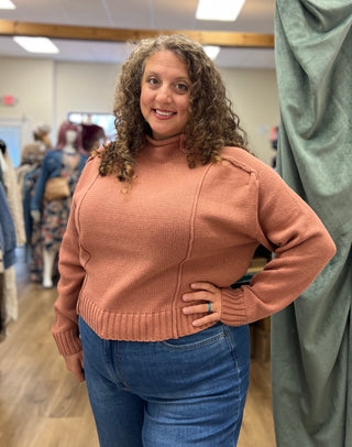 A woman with curly brown hair wearing a dusty rose ribbed knit mock-neck sweater and blue jeans, standing in a clothing store.