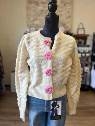 Cream cable-knit cardigan sweater with pink flower-shaped buttons, displayed on a black mannequin torso with blue jeans in a retail store setting.
