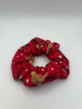 A red fabric hair scrunchie featuring a holiday print of brown reindeer and white stars, displayed on a neutral light gray background.