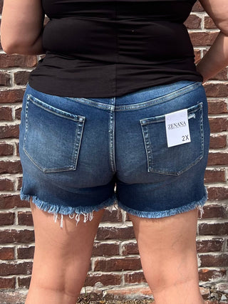 Rear view of a person wearing blue denim cutoff shorts with a frayed hem and a white Zenana 2X size tag on the back pocket, standing against a brick wall.