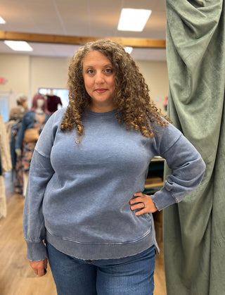 A woman with curly brown hair wearing a washed slate blue crewneck sweatshirt and blue jeans, standing in a clothing store.