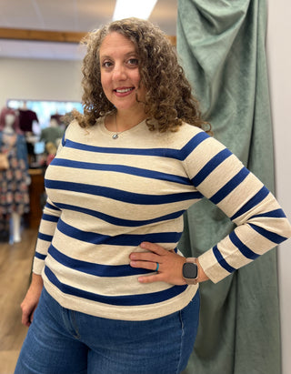 A woman with curly brown hair wearing a cream and navy blue horizontally striped long-sleeve knit top and blue jeans, standing in a boutique setting.