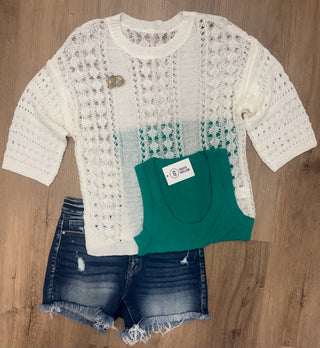 A three-piece women's casual outfit set laid flat on a wooden floor, featuring a cream open-knit long-sleeve top, a folded green tank top, and distressed denim shorts.