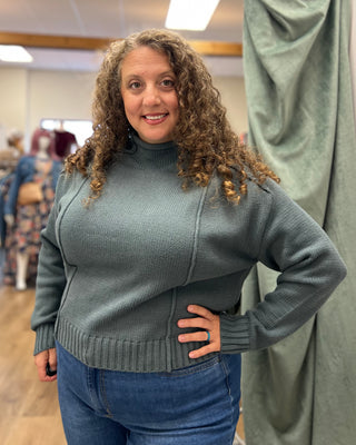 A woman with curly brown hair wearing a slate blue-gray ribbed turtleneck sweater and blue jeans, standing in a boutique with a green curtain behind her.