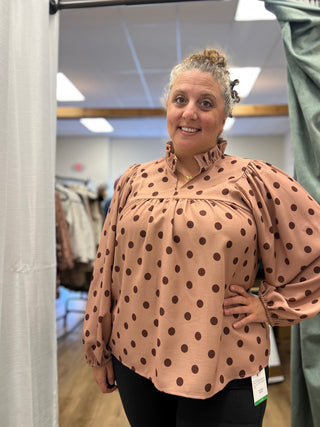 A woman modeling a peach-colored long-sleeve blouse with dark brown polka dots, standing in a fitting room or boutique.