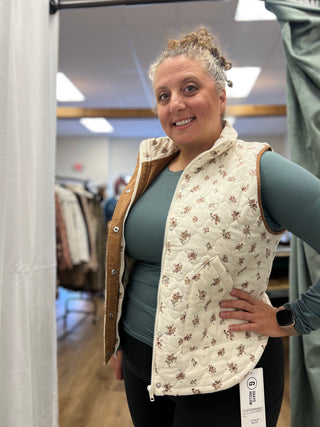 A woman modeling a cream quilted vest with a brown floral pattern and tan corduroy trim, worn over a slate blue long-sleeve shirt in a boutique setting.