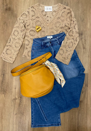 A styled flat-lay outfit on a wood floor featuring a beige crochet cardigan, blue jeans, a mustard yellow leather shoulder bag, and a patterned scarf.