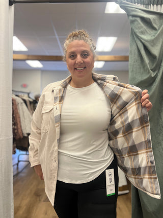 A woman in a fitting room wearing a cream ribbed button-up shirt with a plaid lining, layered over a white t-shirt and black leggings. She is holding open the shirt to show the interior plaid pattern, with a clothing store interior visible in the background.