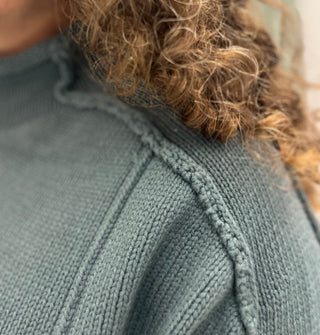 Close-up of a person wearing a textured knit sweater in a muted sage green color, with curly brown hair visible at the neckline.