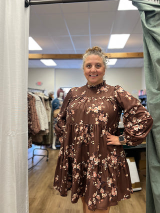A woman modeling a brown floral tiered dress with long balloon sleeves in a boutique fitting room setting.