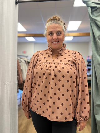 A woman wearing a peach-colored long-sleeve blouse with dark brown polka dots, standing in a fitting room.