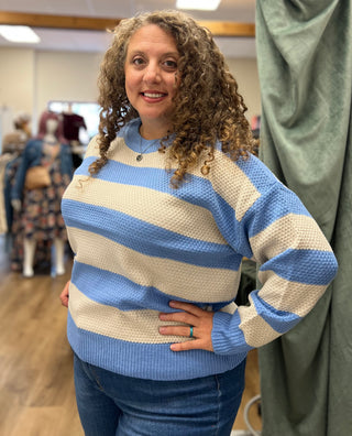 A woman with curly brown hair wearing a blue and cream waffle-knit striped sweater and blue jeans, standing in a clothing boutique.
