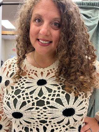 A smiling woman with curly brown hair wearing a cream crocheted top with a black sunflower pattern, photographed in an indoor setting.