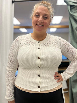 A woman wearing a cream-colored ribbed knit cardigan with lace detailing on the yoke and sleeves, featuring dark brown buttons, shown in an indoor setting.