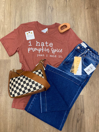 A styled flat-lay outfit on a wood floor featuring a rust-colored graphic t-shirt with the text 'i hate pumpkin spice yeah i said it.', folded blue jeans, and a brown and black-and-white checkered shoulder bag.