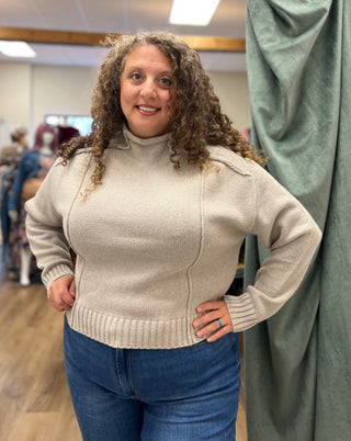 A woman with curly brown hair wearing a beige ribbed mock-neck sweater and blue jeans, standing in a fitting room with a green curtain.