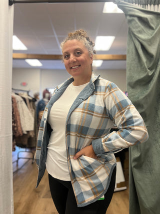 A woman modeling a blue, white, and tan plaid button-up shirt over a white t-shirt and black pants, standing in a fitting room.