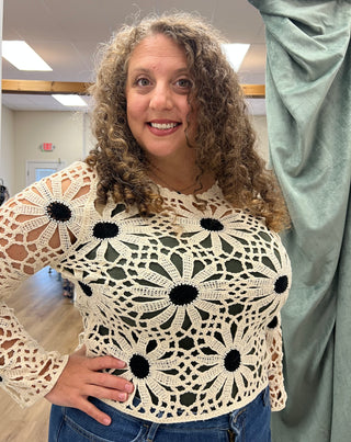 A woman with curly brown hair wearing a cream crochet top with black flower centers, standing in an indoor room with a green curtain on the right.