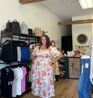 A woman modeling a white puff-sleeve maxi dress with a vibrant orange, pink, and green floral print, standing inside a clothing boutique.