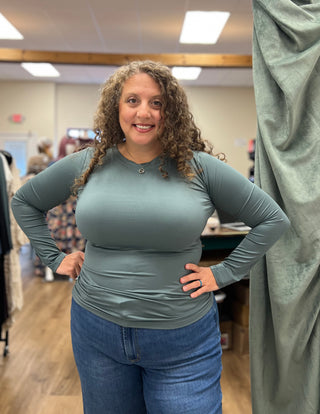 A woman with curly brown hair wearing a fitted sage green long-sleeve top and blue jeans, standing in a retail store with her hands on her hips.