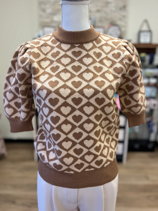 A brown and cream knit sweater with a heart pattern, displayed on a mannequin torso in a softly blurred indoor setting.