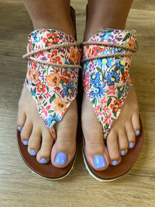 A pair of thong sandals being worn on feet, shown from the top on a wood floor. The sandals have brown platform soles and floral fabric straps with a rope detail across the instep.
