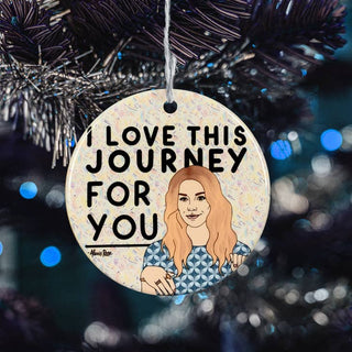 A round ceramic Christmas ornament hanging from a black tinsel Christmas tree. The ornament features a cream background with colorful confetti, the text 'I LOVE THIS JOURNEY FOR YOU' in black, and an illustration of a woman with long orange hair wearing a blue geometric patterned top.