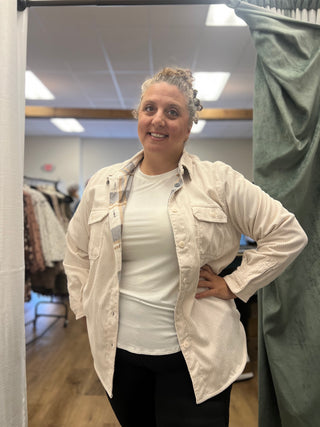 A woman posing in a fitting room wearing a light beige textured button-up shirt over a white t-shirt and black pants.