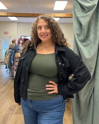 A woman with curly brown hair stands in a clothing store wearing an olive green tank top, a black jacket, and blue jeans, posing in front of a gray curtain.