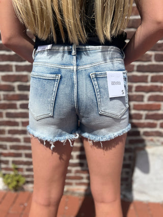 A person shown from the back wearing light-wash frayed denim shorts with a white paper tag attached to the back pocket, standing in front of a brick wall.