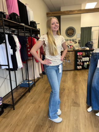 A woman modeling light-wash blue flare jeans and a cream floral short-sleeve top inside a clothing boutique with wood floors and racks of garments.