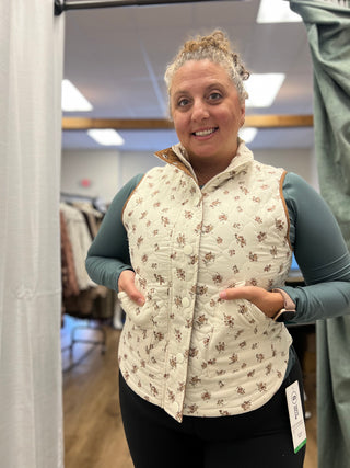 A woman modeling a cream quilted vest with a brown floral pattern, worn over a teal long-sleeve shirt and black pants in a retail fitting room.